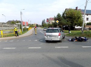 zdarzenie drogowe w Kurzętniku- z prawej strony drogi widać przewrócony na bok motorower, obok niego stoi samochód marki Opel. Po prawej stronie jezdni stoi policjant wykonujący oględziny