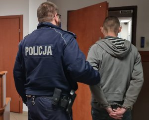 na zdjęciu widać policjanta doprowadzającego zatrzymanego, który na ręce z tyłu ma założone kajdanki