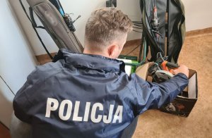 na zdjęciu widać na pierwszym planie policjanta w kurtce z napisem POLICJA. Za nim w tle widać zabezpieczone przez funkcjonariuszy wędki i pilarkę elektryczną pochodzące z kradzieży