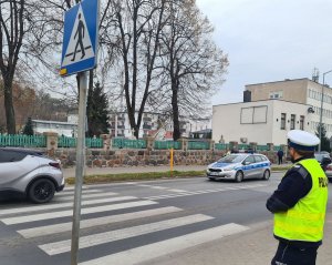 zdjęcie przedstawia policjanta, który stoi w pobliżu przejścia dla pieszych nadzorując ruch. W tle widać cmentarz i przejeżdżający policyjny radiowóz