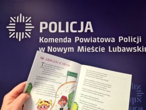 na zdjęciu widać książeczkę &quot;Na ratunek Poli&quot; a w tle baner Komendy Powiatowej Policji w Nowym Mieście Lubawskim