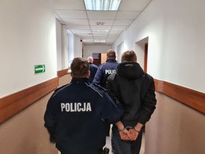 na zdjęciu widać mężczyznę w kajdankach, którego prowadzą korytarzem policjanci