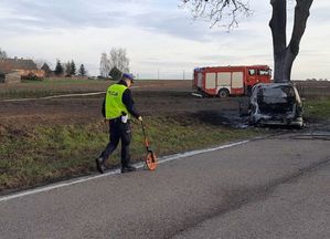 policjant robi oględziny miejsca wypadku drogowego, w tle spalony samochód a za nim wóz strażacki